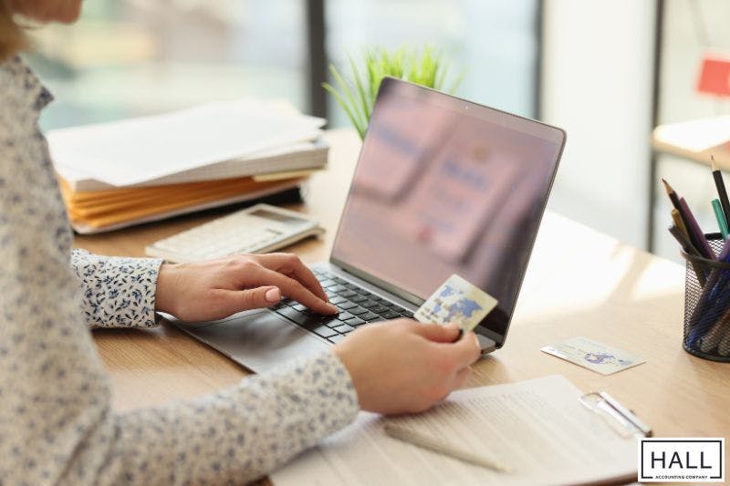 A person sits at a desk using a laptop, holding a credit card while managing financial documents and preparing for tax planning.