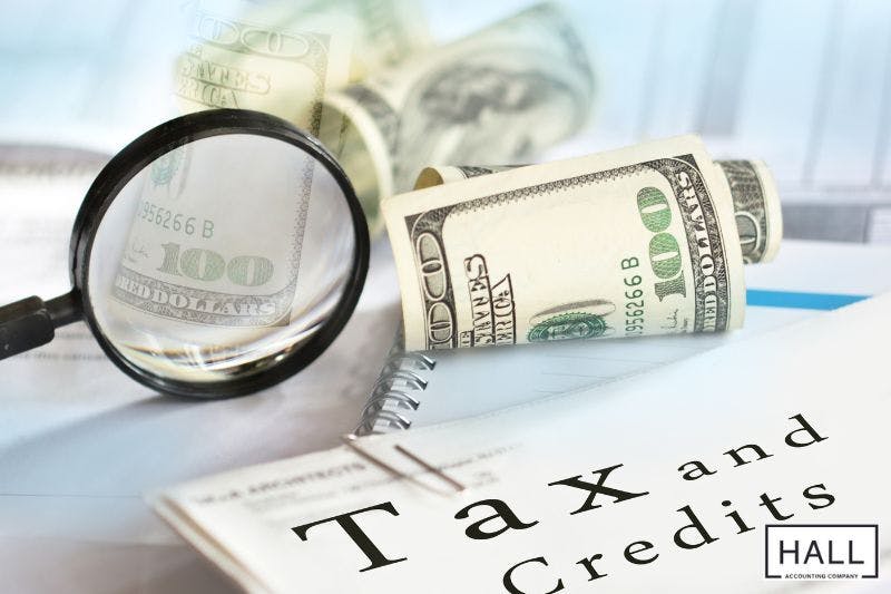 A magnifying glass focuses on a hundred-dollar bill, with additional cash, documents, and a booklet titled 'Tax and Credits' in the background representing the concept of asset exemptions that can protect your tax refund