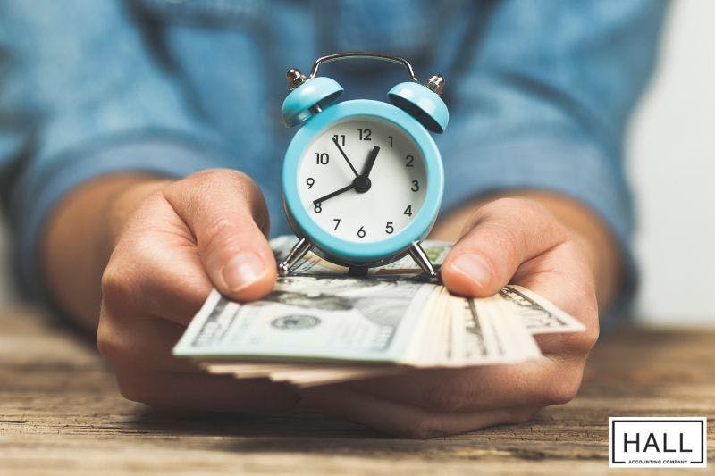 A person holds a stack of dollar bills with a small blue alarm clock resting on top, symbolizing managing time and earnings key considerations for part-time workers looking to optimize their tax strategy.