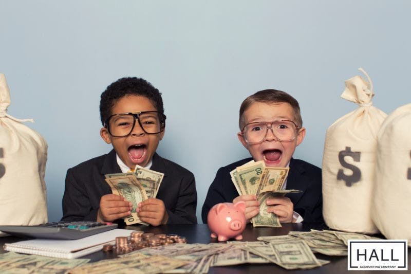 Two young children sit at a table with large money bags, stacks of cash, coins, and a piggy bank