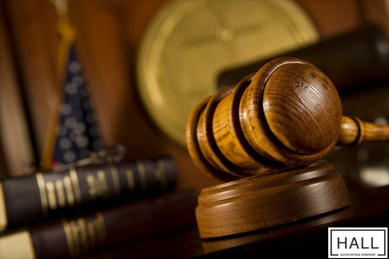 A wooden gavel rests on a sound block in a courtroom setting and behind the gavel are stacks of law books, a flag, and a seal