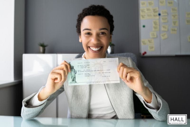 A smiling businesswoman sits at her desk, proudly holding up a tax refund cheque in both hands.