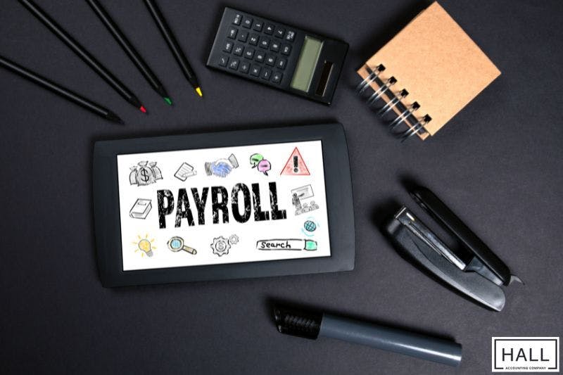 A tablet displaying the word "PAYROLL" surrounded by office supplies like pencils, a calculator, a notebook, and a stapler illustrating payroll managment
